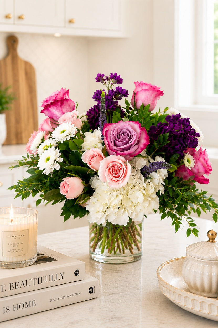 Bouquet of pink, purple, and white flowers in a glass vase on a table with books and a candle.