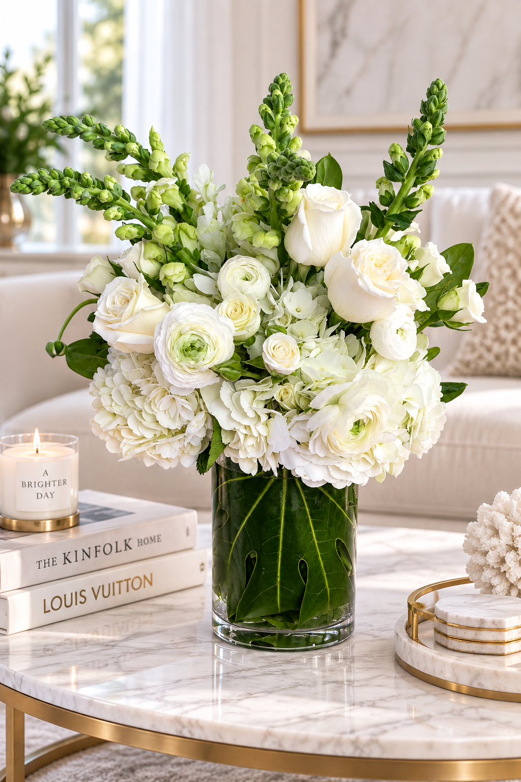 A large floral arrangement of white roses, ranunculus, hydrangeas, and green foliage in a clear glass vase, styled on a marble coffee table in a bright living room setting.