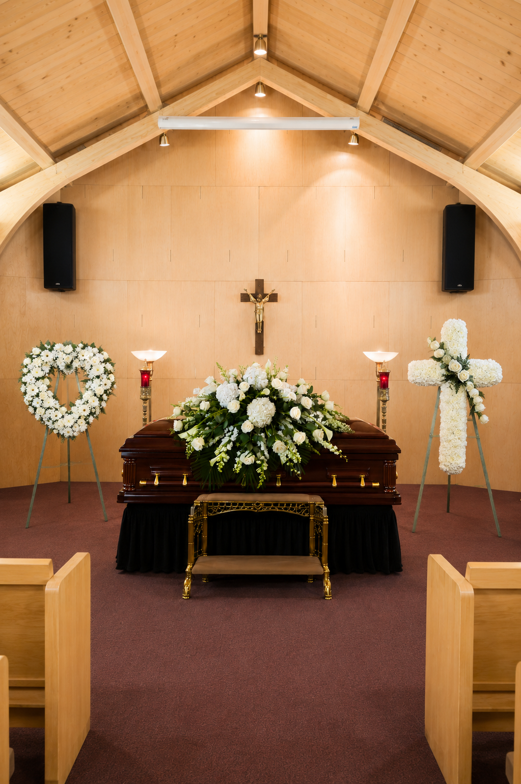 A funeral chapel interior with a dark wooden casket draped in black cloth and decorated with a large white floral arrangement. A gold-framed cross stand sits in front of the casket, with white floral wreaths on stands on both sides.