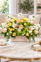 A large floral arrangement in a rectangular wooden box, featuring yellow roses, peach roses, white flowers, and green foliage, displayed on a marble coffee table in a bright living room setting.