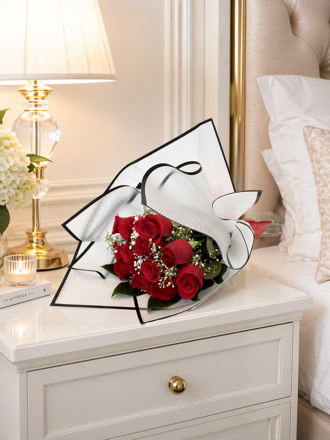 Bouquet of red roses in a decorative box on a nightstand with a lamp and bed in the background.