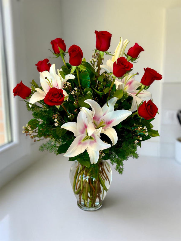 Bouquet of red roses and white lilies in a clear vase on a white surface with a window in the background.